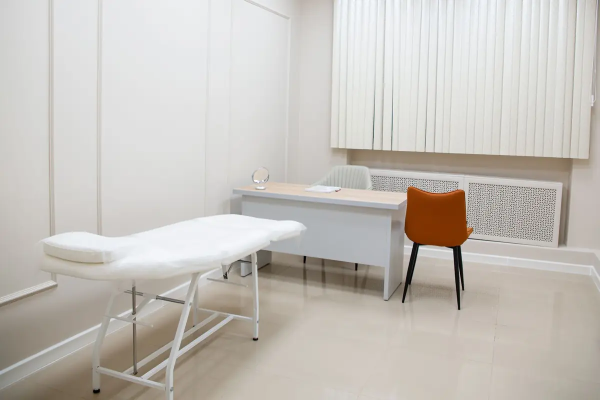 An examination room with a treatment couch, desk and chair beside a window